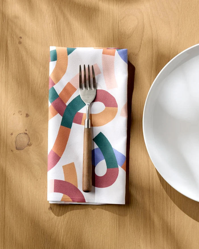 The Claire Iglesias Rainbow Squiggles reusable napkin lies beside a white plate on a light wooden table, with a metal fork featuring a wooden handle resting on it as sunlight casts soft shadows across the scene.