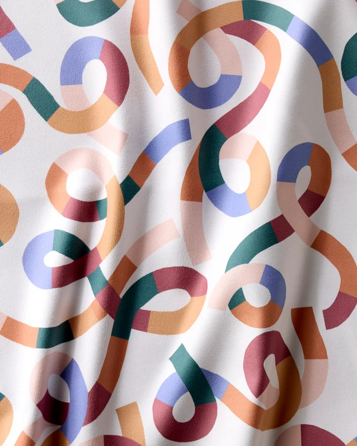 Rainbow Squiggles by Claire Iglesias features vibrant ribbon-like loops in red, orange, green, blue, and purple on white with subtle fabric folds—ideal for a lively set of reusable cloth napkins.