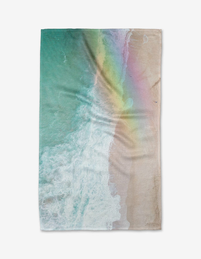 The Rainbow Wave blanket by Kait Hanson is crafted from recycled materials and showcases an aerial print of ocean waves meeting sandy beach in soft blue, turquoise, beige, and pastel rainbow hues.