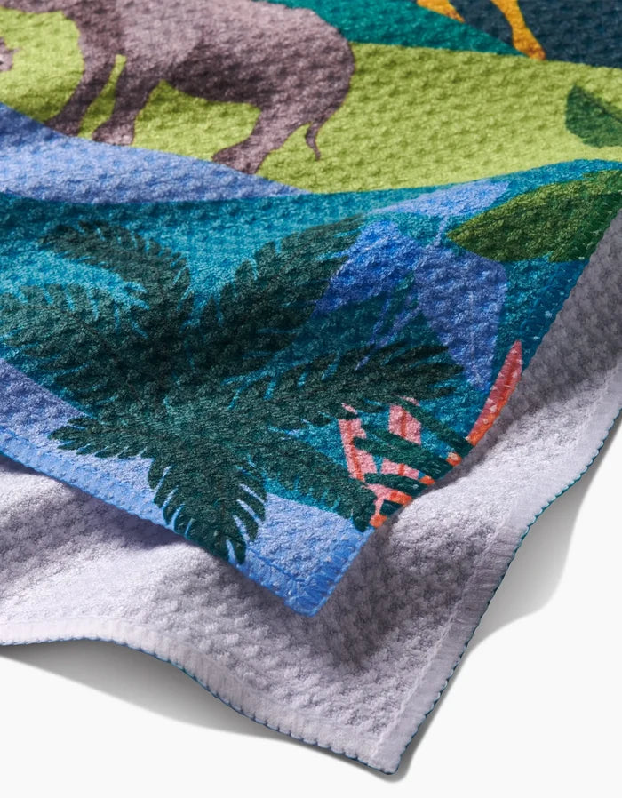 A close-up of Ceyda Alasar’s “Rainforest” bar towel, made from recycled materials, shows its colorful safari design with a rhinoceros, green leaves, tropical plants, and its light gray underside.