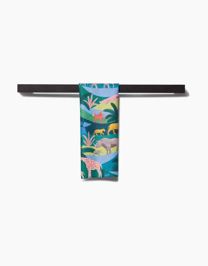The Rainforest bar towel by Ceyda Alasar, featuring vibrant lions, elephants, and giraffes on recycled fabric, is draped over a sleek black horizontal bar against a white background.