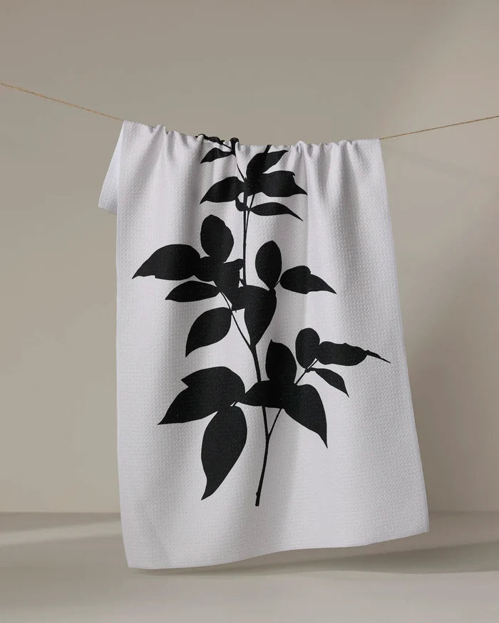 A PrintedPlant "Raspberry" white tea towel featuring a bold black botanical silhouette hangs on a thin clothesline against a neutral background.
