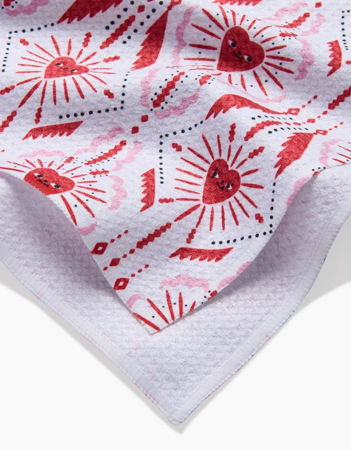 The Ray of Love tea towel by Roxy Marj features a red heart and sunburst design with black and red decorative dots on a white background. Its textured edge is visible in the foreground.
