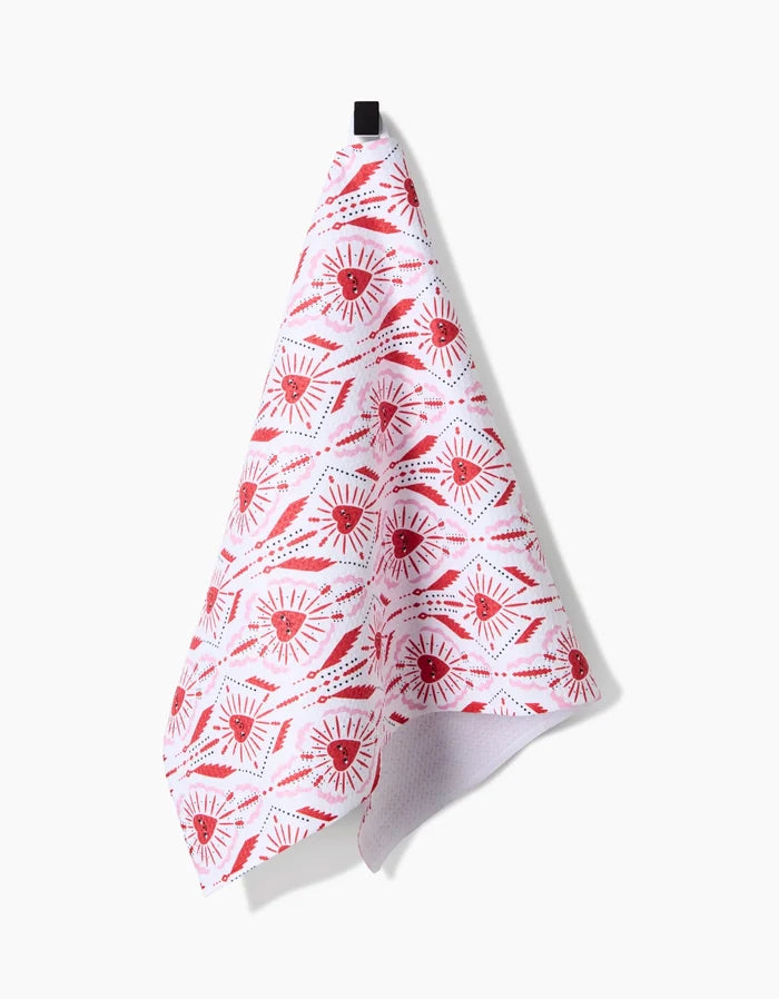 The Ray of Love tea towel by Roxy Marj, featuring red stylized eyes and sunbursts, hangs from a black hook against a plain white background.