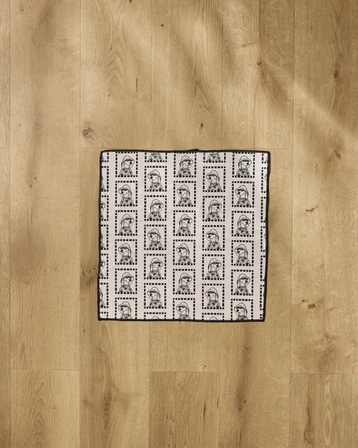 The Geometry "Ready For Summer Doggy" is a square recycled PET towel featuring a grid of postage stamp designs with a woman’s portrait, shown laid flat on a wooden floor.