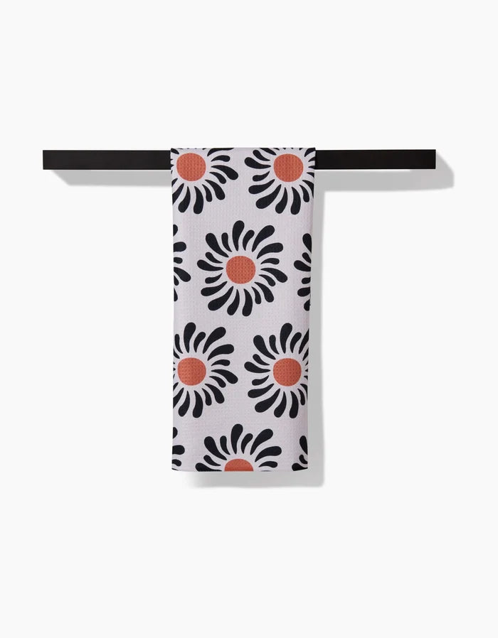 The Realm Refillery eco-friendly white tea towel, featuring a black petal and red center floral design, hangs neatly on a black bar against a white background.