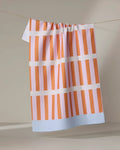 The "Red Glow" tea towel by Happy Hemera, featuring pink and orange stripes and a light blue border, is made from recycled materials and hangs on a clothesline against a neutral backdrop.