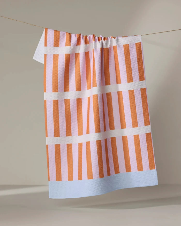 The "Red Glow" tea towel by Happy Hemera, featuring pink and orange stripes and a light blue border, is made from recycled materials and hangs on a clothesline against a neutral backdrop.