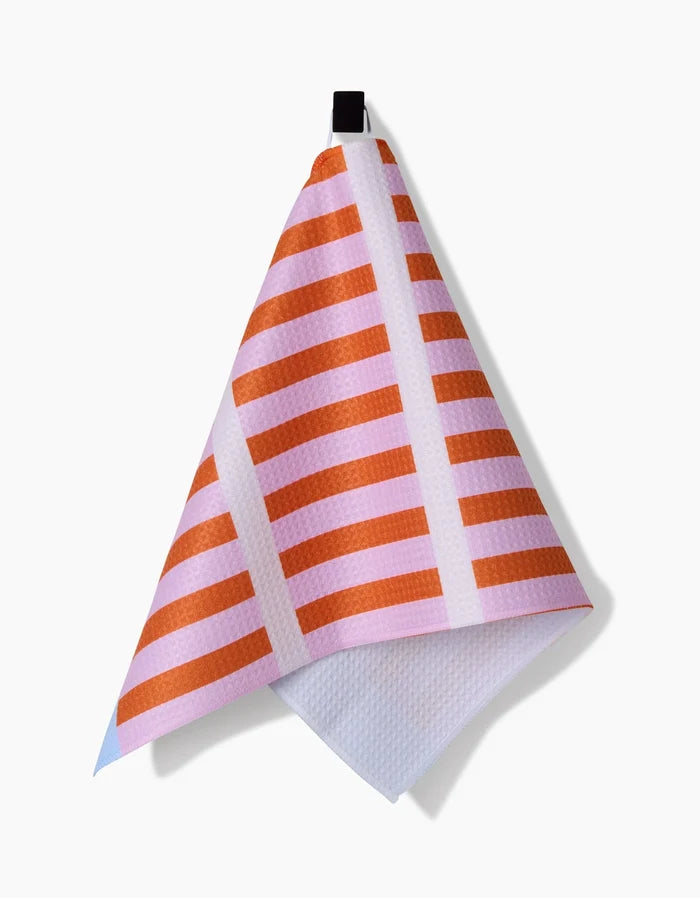The Red Glow Bar Towel by Happy Hemera, crafted from soft Geoweave fabric, features orange horizontal stripes and white lines on a pink background, and hangs from a black loop against a white backdrop.
