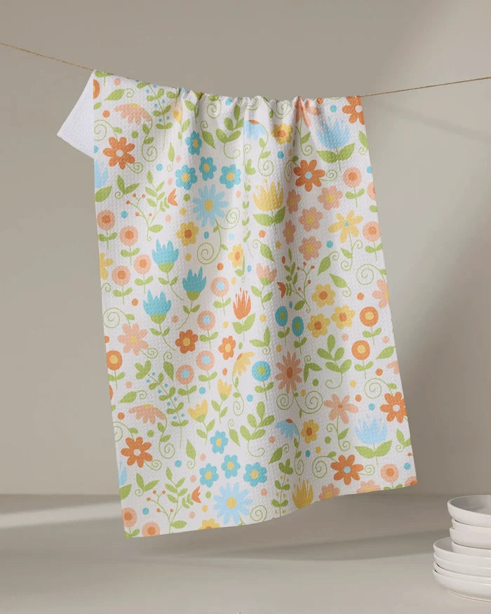 The Mindfulnice Remindful Rememberings tea towel, featuring a vibrant floral pattern in orange, blue, pink, and green on white, hangs on a clothesline with stacked white plates nearby.
