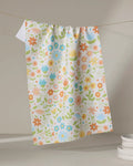 The Mindfulnice Remindful Rememberings tea towel, featuring a vibrant floral pattern in orange, blue, pink, and green on white, hangs on a clothesline with stacked white plates nearby.