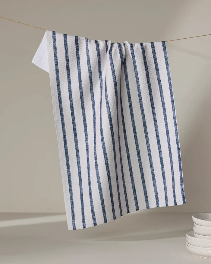 The Becki Owens Ren Blue tea towel, featuring vertical blue stripes and made from recycled materials, hangs on a string against a neutral background. A stack of white dishes is partially visible in the lower right corner.