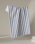 The Becki Owens Ren Blue tea towel, featuring vertical blue stripes and made from recycled materials, hangs on a string against a neutral background. A stack of white dishes is partially visible in the lower right corner.