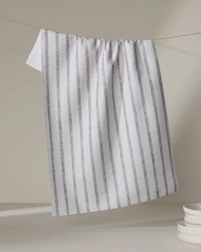 The Ren Grey tea towel by Becki Owens, featuring white fabric with vertical gray stripes and made from recycled materials, hangs on a string with part draping down beside a stack of white plates in the neutral background's lower right corner.