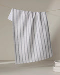 The Ren Grey tea towel by Becki Owens, featuring white fabric with vertical gray stripes and made from recycled materials, hangs on a string with part draping down beside a stack of white plates in the neutral background's lower right corner.