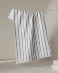 The Becki Owens Ren Sage recycled kitchen towel, white with vertical green stripes, hangs on a string against a light gray backdrop, with a stack of white plates partly visible in the lower right corner.