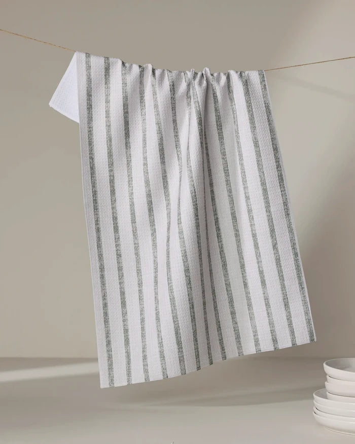 The Becki Owens Ren Sage recycled kitchen towel, white with vertical green stripes, hangs on a string against a light gray backdrop, with a stack of white plates partly visible in the lower right corner.