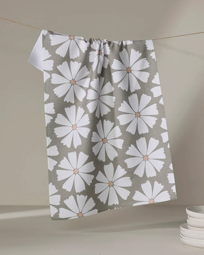 The Hufton Studio Retro Daisy tea towel, made from recycled materials and featuring large white flowers with brown centers, hangs on a line. A stack of white plates sits in the lower right corner against a neutral backdrop.