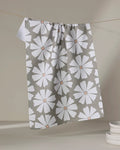 The Hufton Studio Retro Daisy tea towel, made from recycled materials and featuring large white flowers with brown centers, hangs on a line. A stack of white plates sits in the lower right corner against a neutral backdrop.