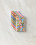Six Retro Garden chocolate bars by Lisa Nelson, each wrapped in pastel floral-patterned, eco-friendly kitchen towels, are fanned out on a marble surface.