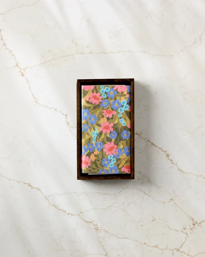 The "Retro Garden" rectangular chocolate bar by Lisa Nelson, wrapped in gold floral paper with pink and blue flowers, sits in a brown box on white marble next to eco-friendly kitchen towels.