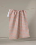 The Retro Geo tea towel by Teri the Butter, made from recycled materials and featuring a pink and white geometric pattern, hangs on a clothesline against a plain beige background.