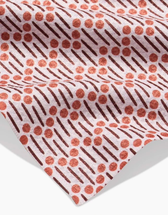 A close-up of Teri the Butter’s Retro Geo Bar Towel on a pink background displays its Geoweave fabric, crafted from recycled materials, with diagonal dark lines and scattered red dots enhanced by soft folds and shadows.