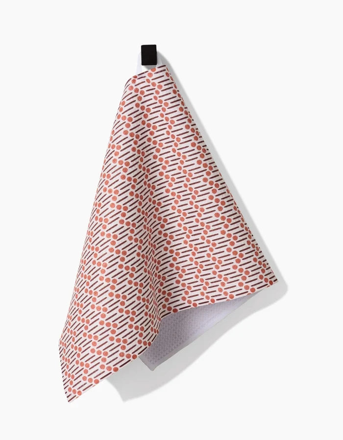 The Retro Geo bar towel by Teri the Butter, made from recycled materials and patterned with small orange dashes, hangs from a black loop against a plain white background.
