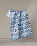 The Retro Rain tea towel by Anette Heiberg, featuring blue and white patterns with black teardrop shapes, hangs from a clothesline against a plain beige background.