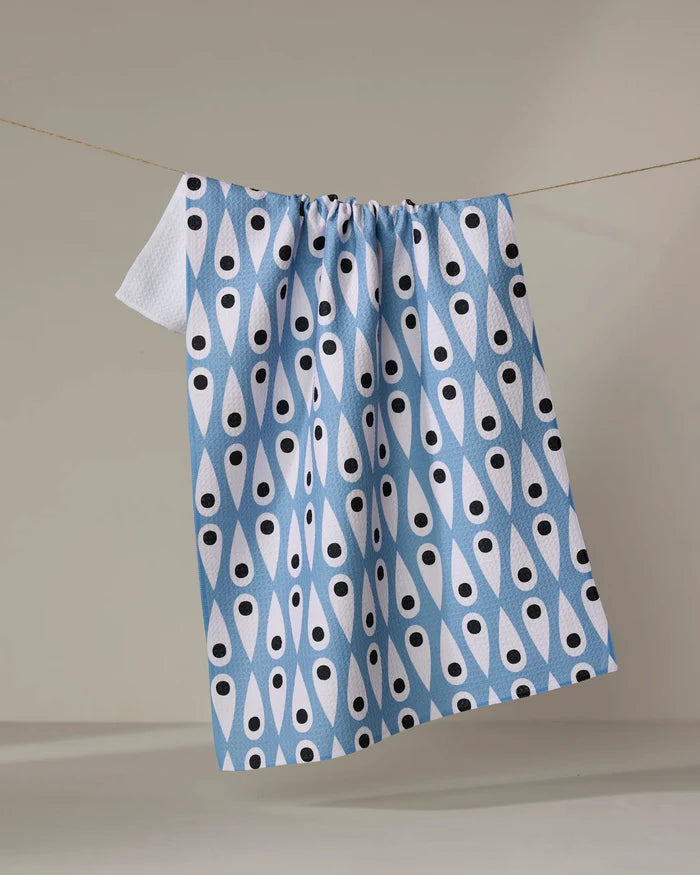 The Retro Rain tea towel by Anette Heiberg, featuring blue and white patterns with black teardrop shapes, hangs from a clothesline against a plain beige background.
