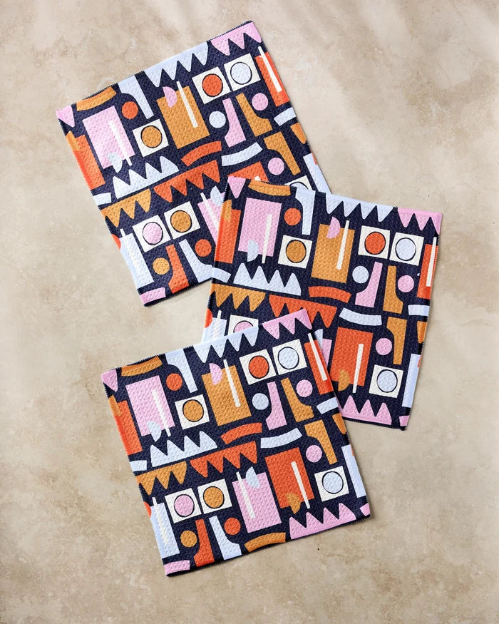 The Claire Iglesias "Retro Shapes Fiesta" set features three rectangular dishcloths with bold geometric patterns in pink, orange, white, and black—super-absorbent and crafted from recycled materials.