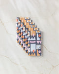 Six Claire Iglesias "Retro Shapes Fiesta" chocolate bars with colorful geometric wrappers, arranged diagonally on white marble veined with tan—evoking an eco-friendly feel inspired by recycled paper towel alternatives.