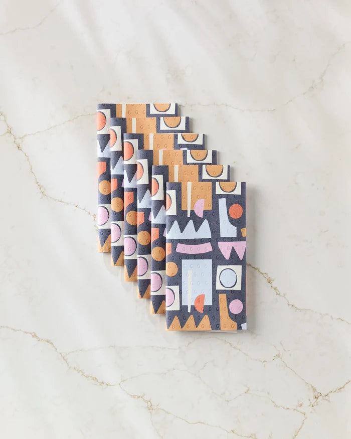 Six Claire Iglesias "Retro Shapes Fiesta" chocolate bars with colorful geometric wrappers, arranged diagonally on white marble veined with tan—evoking an eco-friendly feel inspired by recycled paper towel alternatives.