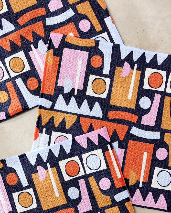 The Retro Shapes Fiesta dishcloth set by Claire Iglesias includes four eco-friendly cloths made from recycled materials, each featuring colorful geometric patterns in orange, pink, blue, red, white, and yellow. Beautifully arranged on a neutral surface.
