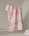 The Retro Vibes tea towel by Marina Solodka, featuring a pink and beige floral pattern and ultra-absorbent fabric, hangs on a clothesline against a neutral background beside a stack of white plates.