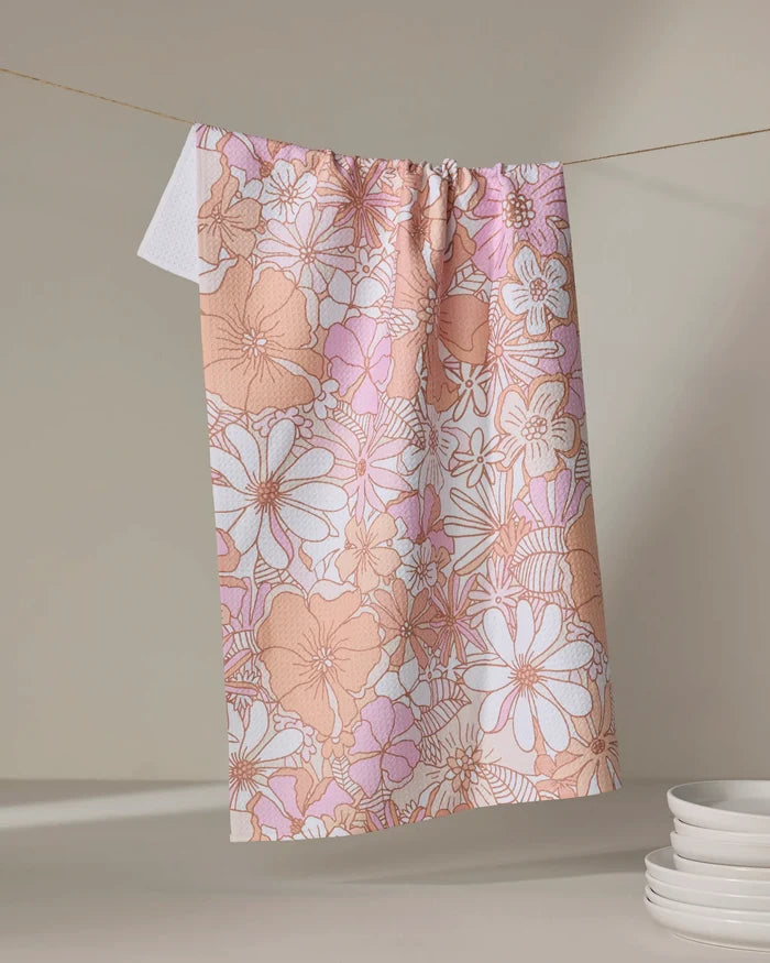 The Retro Vibes tea towel by Marina Solodka, featuring a pink and beige floral pattern and ultra-absorbent fabric, hangs on a clothesline against a neutral background beside a stack of white plates.