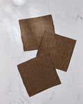 Three Ridge napkins by Geometry, made from recycled materials, feature a black geometric pattern on brown fabric and are arranged on a white marble surface.