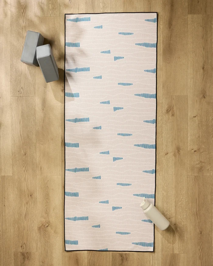 The Twig + Spark Ridgeline yoga mat with blue and white stripes is on a wooden floor, paired with two grey yoga blocks in the top left. A white water bottle and a non-slip towel for stability are at the bottom right.