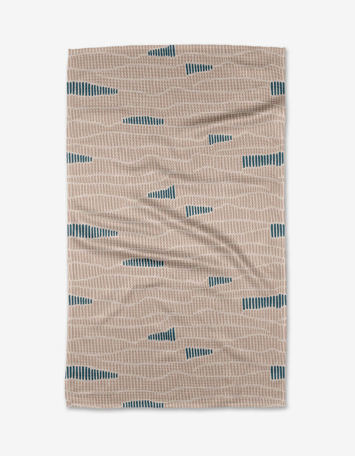 The Ridgeline kitchen towel by Twig + Spark is beige, made from recycled materials, and features an abstract pattern with short light tan lines and small dark teal triangles, shown flat on a white background.