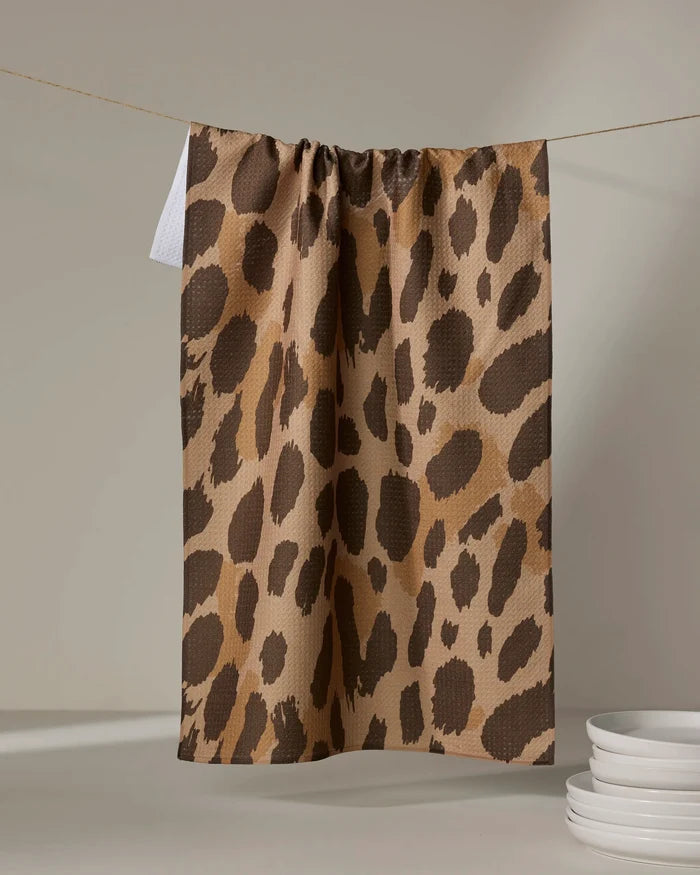 The Geometry Roam absorbent dish towel, featuring a brown and tan leopard print, hangs on a clothesline above a white surface with a small stack of white plates in the lower right corner.