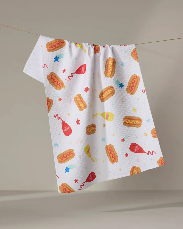 The "Rocket Relish" kitchen towel by Vaishali Design Studio hangs on a clothesline, featuring playful hot dogs, ketchup and mustard bottles, stars, and bursts on white recycled fabric—a festive touch for your kitchen.