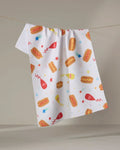 The "Rocket Relish" kitchen towel by Vaishali Design Studio hangs on a clothesline, featuring playful hot dogs, ketchup and mustard bottles, stars, and bursts on white recycled fabric—a festive touch for your kitchen.