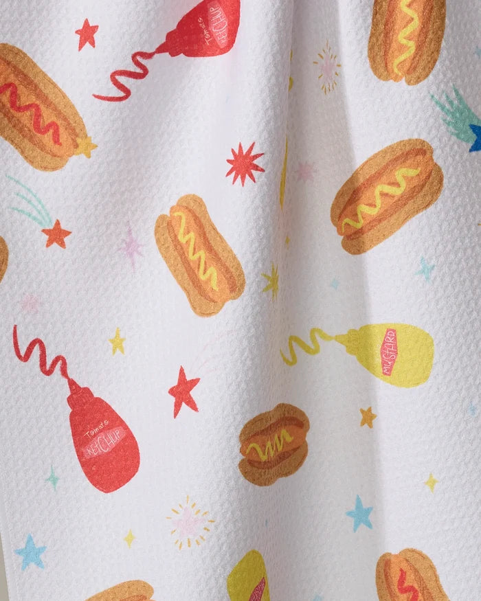 The Rocket Relish kitchen towel from Vaishali Design Studio is a white recycled towel featuring textured fabric and colorful hot dog, ketchup, mustard, shooting star, and starburst illustrations in red, yellow, and blue.
