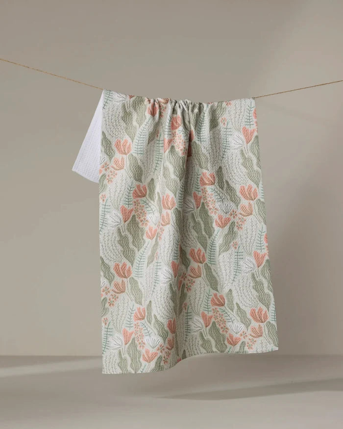 The Amy MacCready Rollie, a recycled kitchen towel with green leaves and peach flowers, hangs from a clothesline against a neutral background.