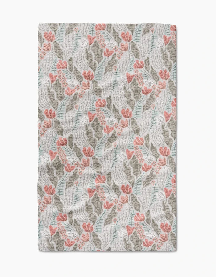 The Rollie blanket by Amy MacCready features a botanical pattern with pink flowers, green leaves, and blue accents on a white background, inspired by Geometry House Tea Towels. Its slightly wrinkled fabric adds extra coziness.