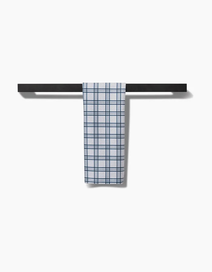 The Roma Bar Towel by Kelsi Fullmer, featuring blue and white checkered Geoweave fabric, hangs neatly on a black horizontal towel bar against a plain white background.