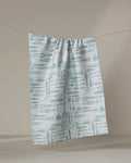 The Romy dish towel from Amy MacCready—a white, absorbent towel with a light blue kitchen utensil pattern—hangs on a string against a plain beige background.