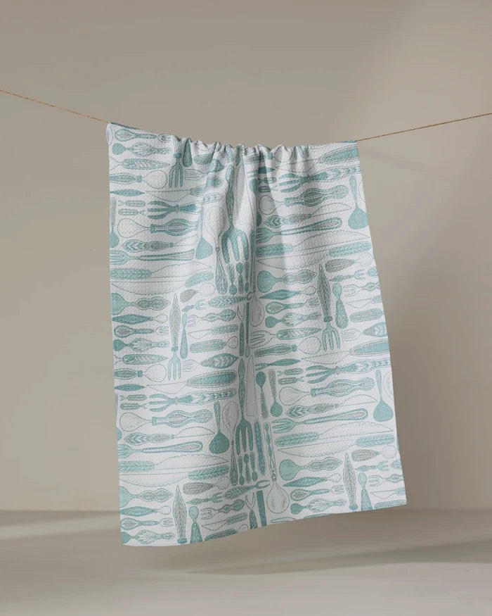 The Romy dish towel from Amy MacCready—a white, absorbent towel with a light blue kitchen utensil pattern—hangs on a string against a plain beige background.
