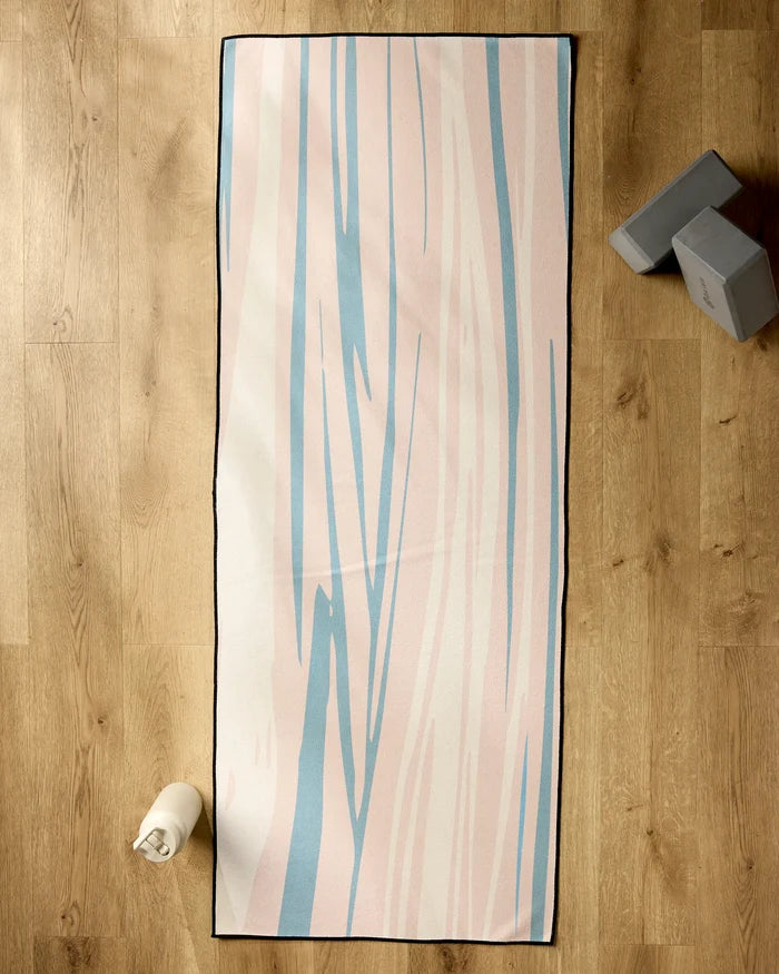 The GEOMETRY Root yoga mat with a pastel pink and blue abstract pattern lies on a wooden floor, paired with a non-slip yoga towel, a water bottle at the lower left corner, and two gray yoga blocks at the upper right.