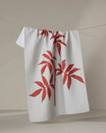 The "Rose" tea towel by PrintedPlant, featuring two large red leafy plant designs, hangs on a clothesline against a plain, light background.